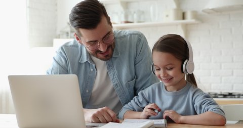 Caring young father helping little primary school learner daughter doing homework, e-learning indoors. Smiling parent explaining education program to small girl in headphones, online study concept.