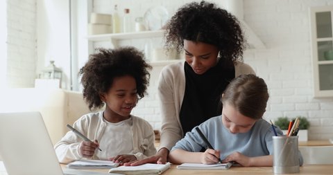 Smiling little cute diverse primary school girls studying with african american female teacher at home. Pretty young biracial foster mother helping two small mixed race daughters with homework.