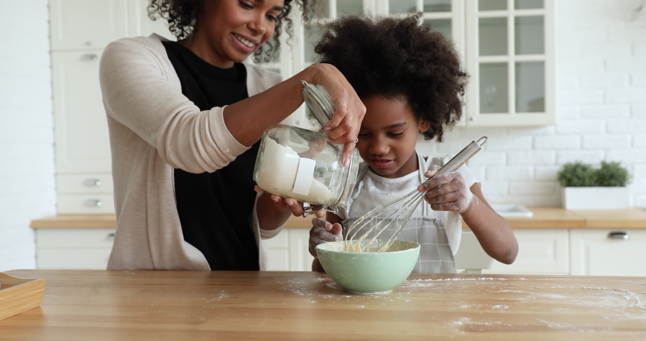 Happy african american young mother adding flour to bowl, teaching little cute biracial child daughter preparing pancakes for weekend breakfast, smiling afro family cooking pastry together in kitchen. Royalty-Free Stock Footage #1056450482