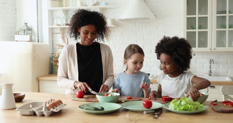 Smiling attractive young afro american mother teaching cooking little multiracial girls children. Happy interracial family preparing healthy lunch, chopping fresh vegetables for salad in kitchen.