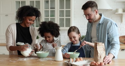 Happy multiracial parents giving culinary class, helping adorable little diverse children daughters mixing flour with milk, making dough for homemade bakery at modern kitchen, cooking skills concept.