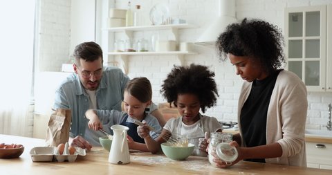 Little cute focused multiracial kids girl wearing aprons, learning making dough for pastry with affectionate loving mixed race parents, interracial family involved in hobby together in modern kitchen.