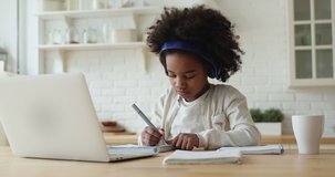 Little adorable focused afro american schoolgirl wearing headphones, studying remote online from home. Concentrated small mixed race kid girl using computer application, learning at video class.