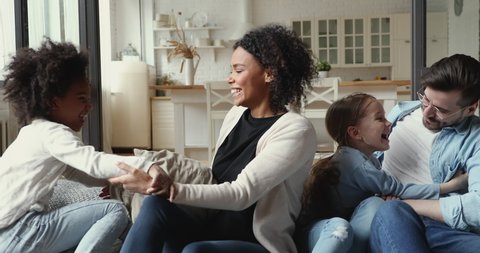 Overjoyed young mixed race couple playing with cute funny little multiracial daughters on sofa indoors. Excited diverse people family enjoying spending weekend time together, having fun at home.