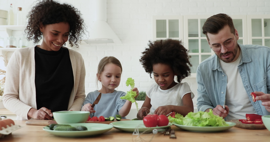 Happy interracial couple teaching little mixed race daughters preparing fresh healthy food in modern kitchen. Smiling multiracial family involved in cooking with adorable small diverse children. Royalty-Free Stock Footage #1056450509