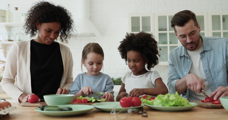 Happy interracial couple teaching little mixed race daughters preparing fresh healthy food in modern kitchen. Smiling multiracial family involved in cooking with adorable small diverse children. | Shutterstock HD Video #1056450509