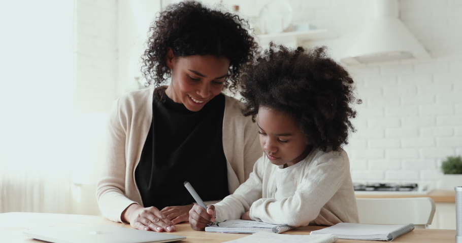 Happy young biracial mother nanny babysitter helping little daughter pupil preparing for test. Focused small afro american primary school girl doing homework with smiling mommy, distant education. Royalty-Free Stock Footage #1056450515