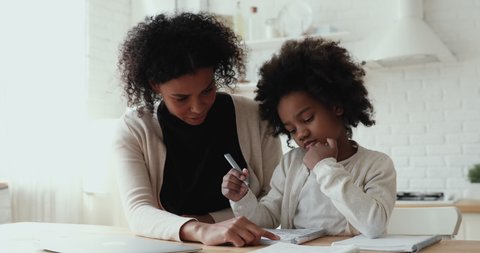 Happy young biracial mother nanny babysitter helping little daughter pupil preparing for test. Focused small afro american primary school girl doing homework with smiling mommy, distant education.