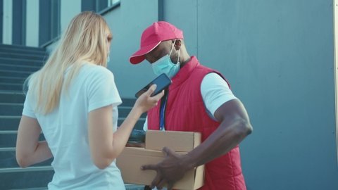 African american man courier wearing a mask delivers a parcel to woman client paying by contactless terminal for parcel transporting payment uniform express order worker outdoors close up slow motion