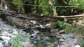 A wooden bridge over a small mountain river with rapids and rocks, among trees entwined with greenery. Taken by a drone rising from the bottom up