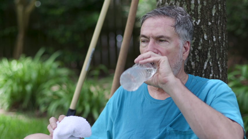 An older man who is sweating profusely from working in his yard shows symptoms of dehydration and heat exhaustion as he sits under a tree and drinks water in an effort to avoid heat stroke. | Shutterstock HD Video #1056452123