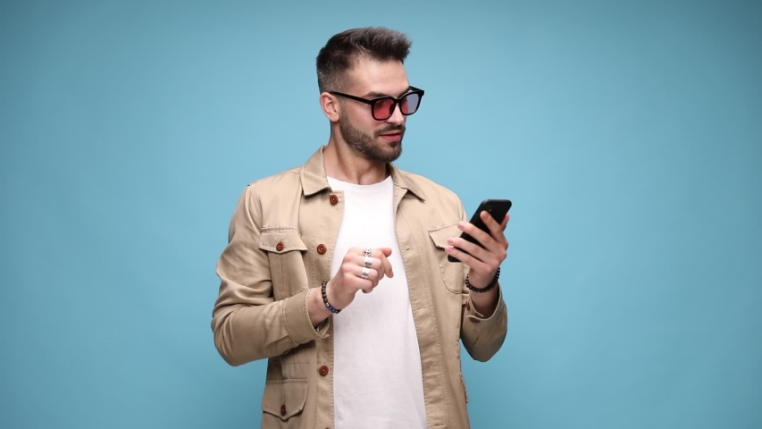 happy casual man in jacket holding phone and reading emails, covering mouth with hand and dancing, pointing finger to phone and making call me sign, moving and gesturing on blue background Royalty-Free Stock Footage #1056458060