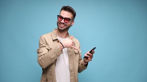 timid young casual guy holding phone and dancing, pointing finger to screen and making call me sign, moving and sticking out tongue on blue background