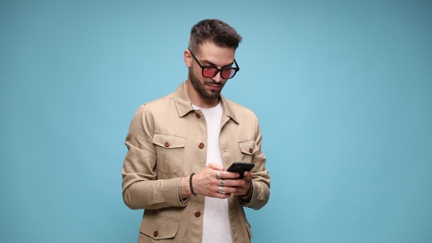 excited young casual man wearing jacket and glasses, looking at the phone and writing messages, pointing two fingers and smiling on blue background Royalty-Free Stock Footage #1056458144