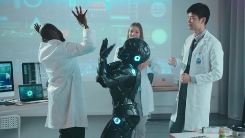 Successful team of multi-ethnic engineers dancing with prototype black cybernetic robot having fun celebration in laboratory. Future is now. Humans and robots. Entertainment.