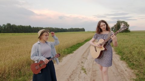 duet of cute cheerful girls. Wheat field road at sunset in summer evening. Guitar and ukulele. Girls sing and laugh. Dynamic camera movement. Slow motion footage