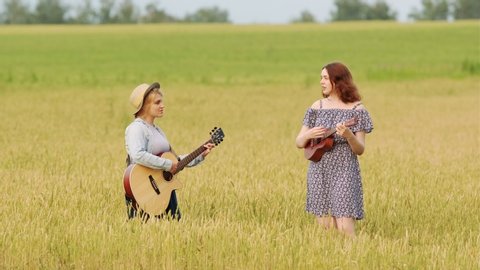 two young beautiful women play country music on guitar and ukulele. Wheat field at sunset on a summer evening