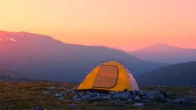 An orange tent stands against the background of a mountain range in the warm sunset light. Hiking in the mountains.