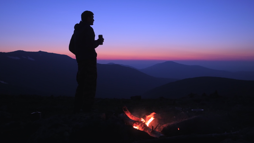 A man drinks a hot drink at night by a campfire in a tourist camp on a mountain pass. A tourist admires the beautiful views of the mountains after sunset. Hiking in the mountains. Royalty-Free Stock Footage #1056470039