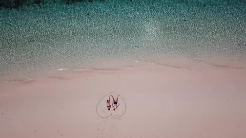 A drone shot of a couple lying on a pink sand beach in a heart drawn on the sand a small island near Maumere, Indonesia. Happy and careless moments. Waves gently washing the shore. Romance and love
