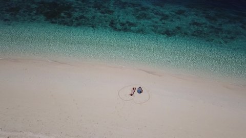 A drone shot of a couple sitting on a pink sand beach in a heart drawn on the sand a small island near Maumere, Indonesia. Happy and careless moments. Waves gently washing the shore. Romance and love

