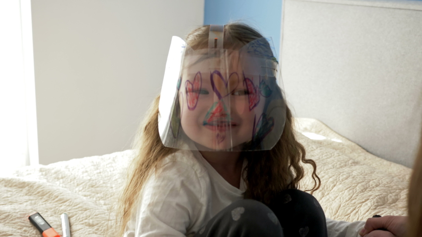 Cheerful little girl in a protective plexiglass mask. There are funny drawings on the mask. Someone is an adult playing with a child.