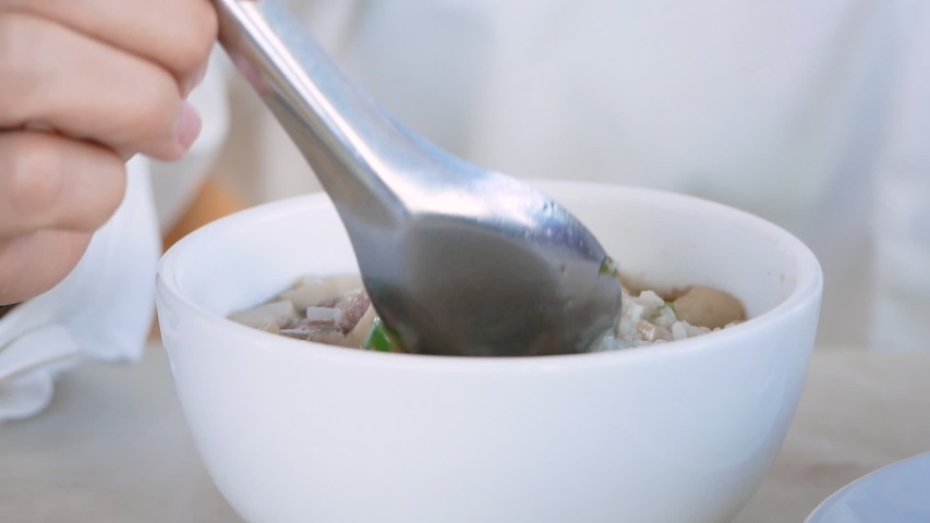 Asian woman eating boiled rice with pork, shiitake mushrooms and vegetables in a white bowl on a table.