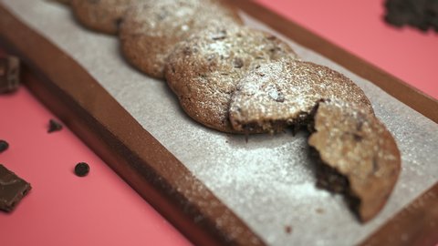 Freshly made Chocolate Chip Cookies on a wooden serving board. Cookies with a glass of fresh milk on the side along with chocolate chips and pieces of chocolate around on a pink background.