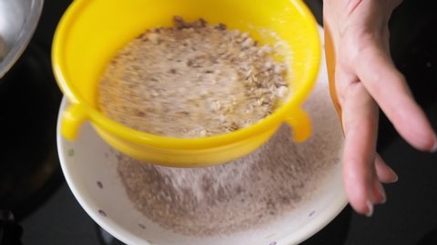 Asian woman sifting flour mixed with cocoa powder through a yellow sieve on the black table.