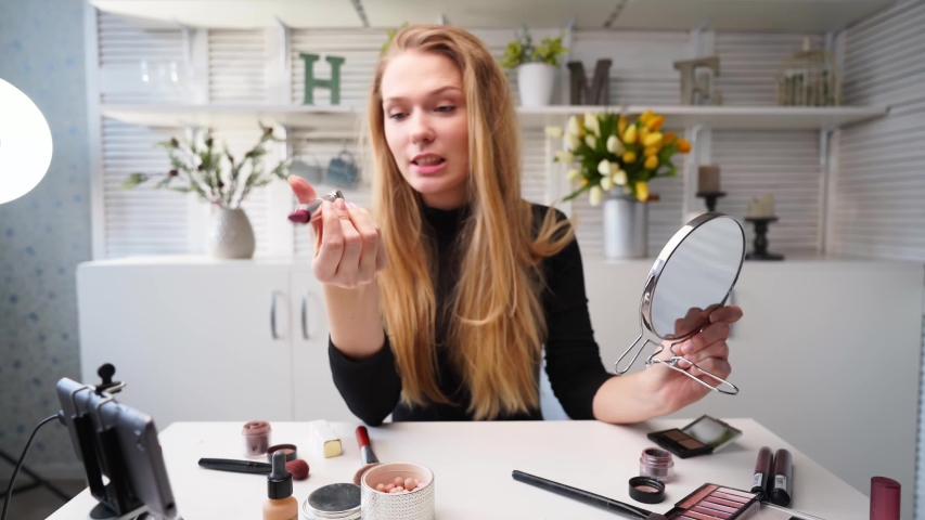 Vlogger female applies lipstick on lips. Beauty blogger woman filming daily makeup routine tutorial at camera on tripod. Influencer blonde girl live streaming cosmetics product review in home studio | Shutterstock HD Video #1056486806