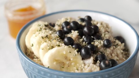 Slow motion adding hemp seeds into oatmeal porridge bowl with bananasand black currants. Healthy breakfast, vegan food concept
