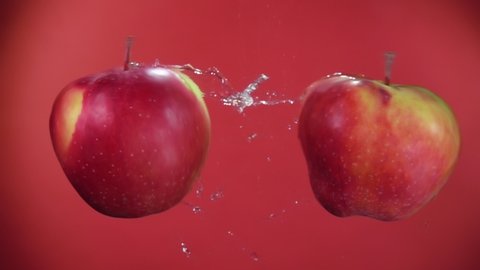 Two large ripe red apples are flying towards each other, colliding on the red background and raising splashes of water in slow motion