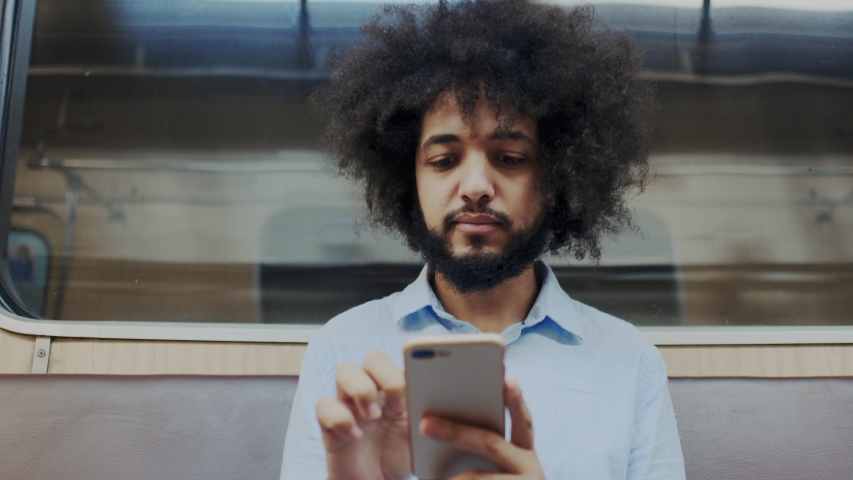 Portrait of Young Stylish Hispanic Man Using Cellular Smartphone in Subway. Curly Man Sitting in Train Wagon at Night and Touches Screen of Mobile Phone. Royalty-Free Stock Footage #1056502472