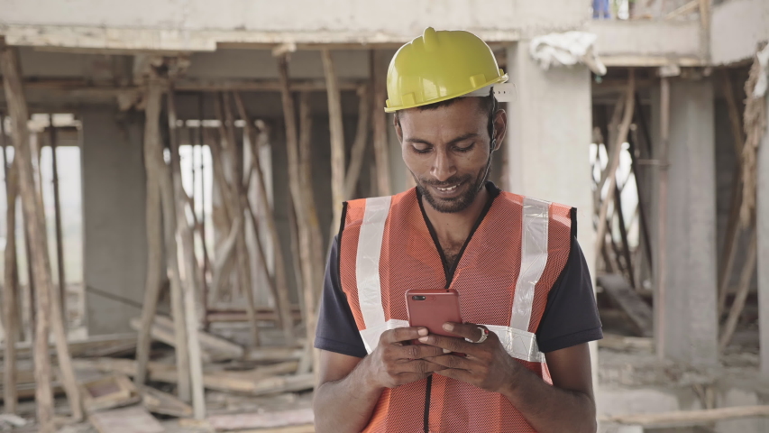 A shot of a man/ male migrant worker or daily wager wearing safety jacket and hard hat standing at under construction building site using mobile phone or smartphone to type a text message | Shutterstock HD Video #1056502826