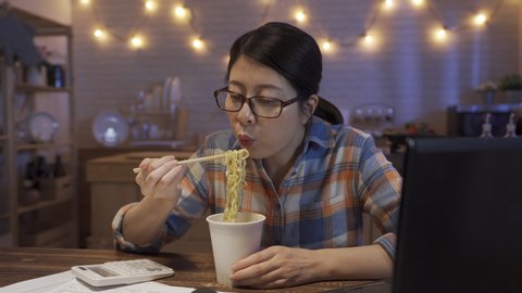 beautiful young asian japanese woman eating instant noodles with chopsticks while working on laptop computer overtime at home kitchen. girl worker feel hungry and enjoy fast food late night meal