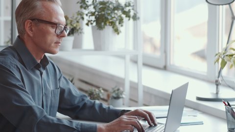 Middle-aged caucasian businessman in shirt and glasses typing on laptop while working at desk in the office