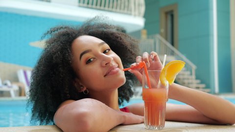 African american woman in red swimsuit drinking non-alcoholic orange cocktail from straw, relaxation sitting in pool water, smiling at camera. Ethnic Girl enjoying vacation at luxury hotel in heat