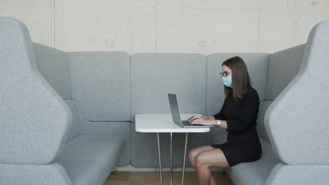 Woman in mask working on laptop in cubicle and social distancing