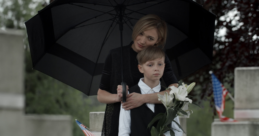 Sad woman hugging and leaned to his teen kid while standing under umbrella at cemetery.Widow and young boy with white flowers near gravestone with american flag.Concept of memorial day. Royalty-Free Stock Footage #1056512204