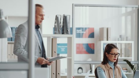 Businessman walking to female colleague in the office, leaning on desk, asking her about document on clipboard and discussing project on laptop during workday