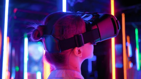 VR, futuristic, retrowave, immersive, entertainment concept. Woman using virtual reality headset and looking around at interactive technology exhibition with colorful illumination - back view