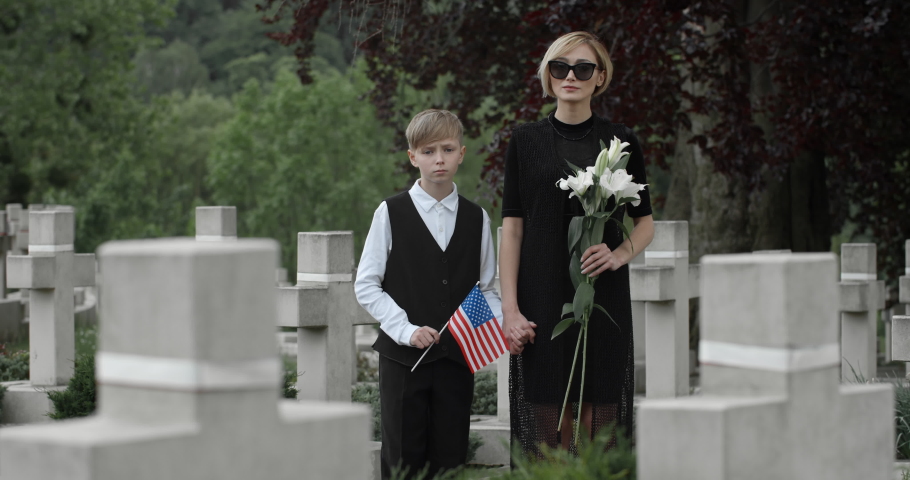Portrait of kid holding american flag and young woman with flower looking to camera. Woman in dark clothes holding her son hand while standing at cemetery. Concept of memorial day.