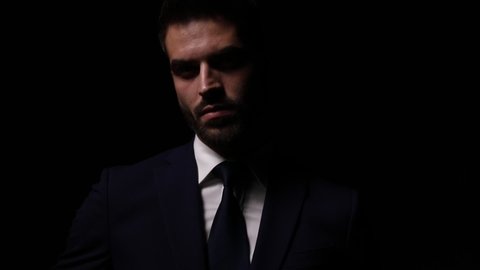 attractive young fashion model in suit, standing in a shadow light and fixing tie, posing and crossing arms on dark background
