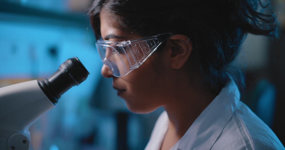 Female research scientist looking into microscope, wearing safety glasses.Blue lighting in a dark lab room.Close up, slider, shot with BMPCC 4K.Biochemistry, pharmaceutical medicine, science concept
 | Shutterstock HD Video #1056523148