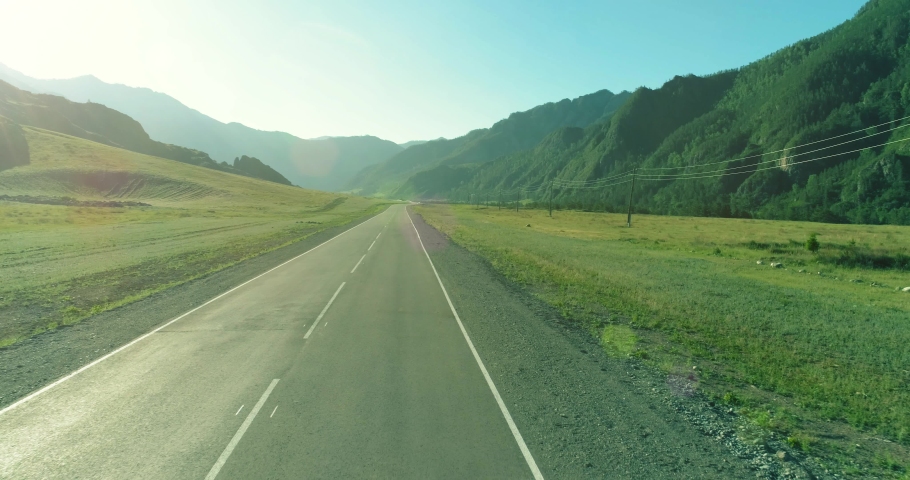 Aerial low air flight over mountain perfect asphalt highway road and meadow at sunny summer morning. Near green trees. Fast horizontal movement with sun rays.