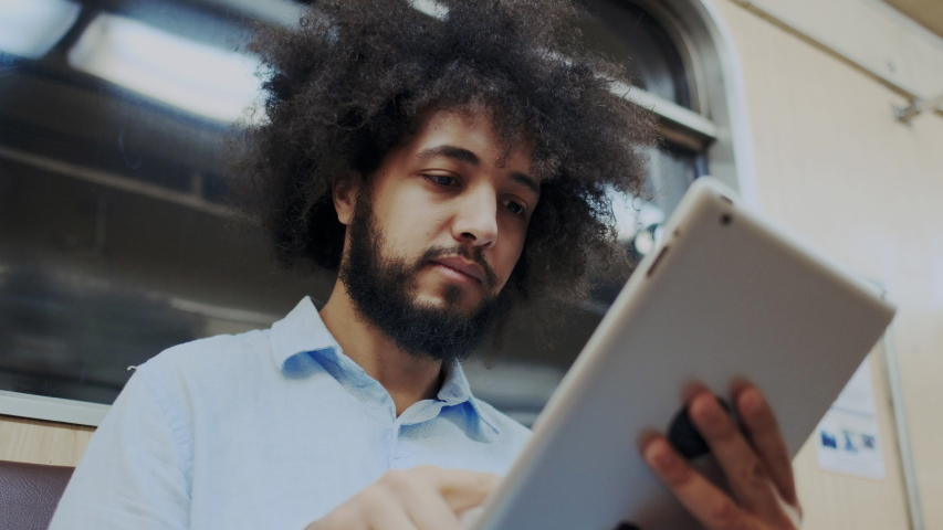 Close-Up Portrait of Young Stylish Hispanic Man Using Cellular Mobile Device in Subway. Curly Man Sitting in Train, Touches Screen of Tablet PC with Fingers. Remote Work, Browsing Internet, Learning. Royalty-Free Stock Footage #1056532040