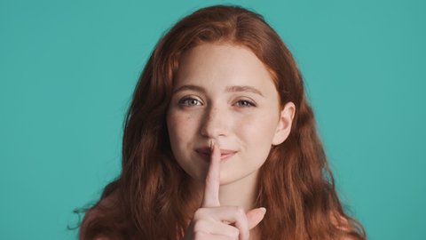 Portrait of redhead woman telling secret showing silence gesture on camera over colorful background. Keep secret expression