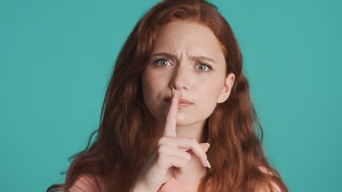 Attractive redhead woman seriously showing silence gesture on camera over blue background. Don't tell expression