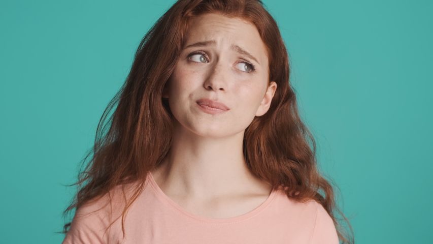 Beautiful worried redhead woman shyly looking in camera and smiling over colorful background. Face expression | Shutterstock HD Video #1056532256