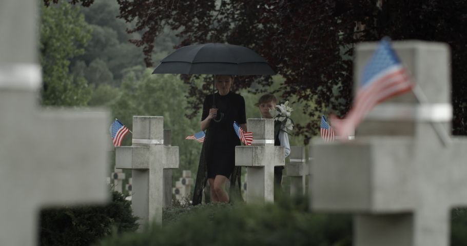Mother and her teen son under umbrella putting flowers at cemetery. Widow and child walking and stopping near gravestone. Concept of memorial day. American flags on stone crosses. Royalty-Free Stock Footage #1056537314